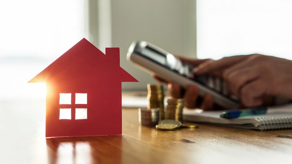 Rotes Haus-Symbol steht auf einem Tisch neben Münzstapeln; im Hintergrund bedient eine Person einen Taschenrechner und macht Notizen zur Finanzierung.